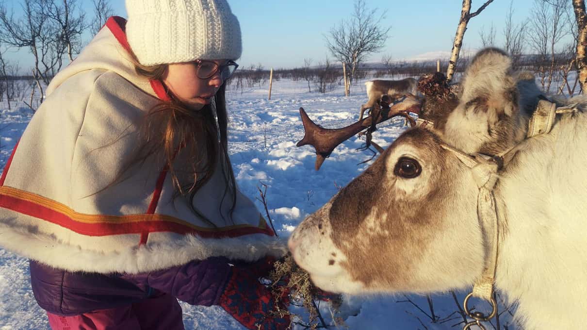 A child in a Sámi-patterned cape feeding a reindeer by hand in the snow