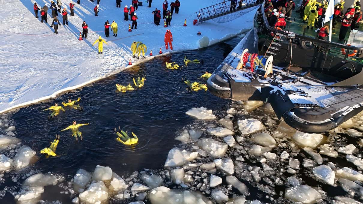 Floaters in yellow survival suits lying in dark water among ice chunks beside the icebreaker