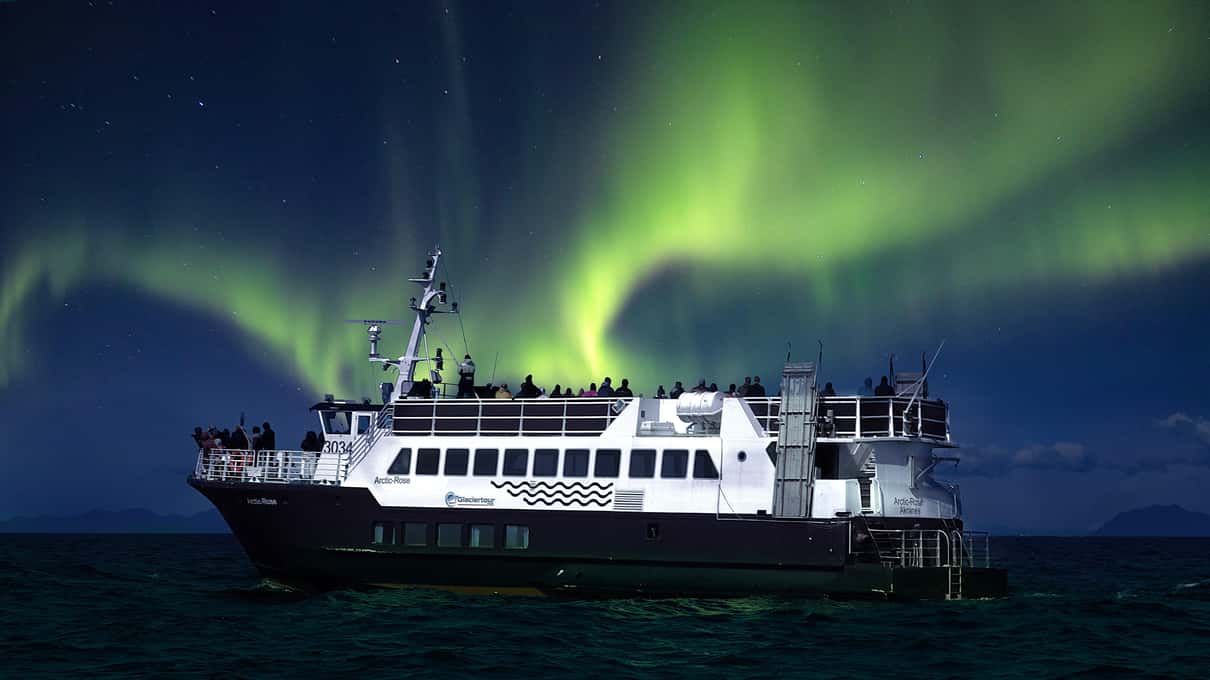A two-deck white cruise boat on black water under swirling green northern lights