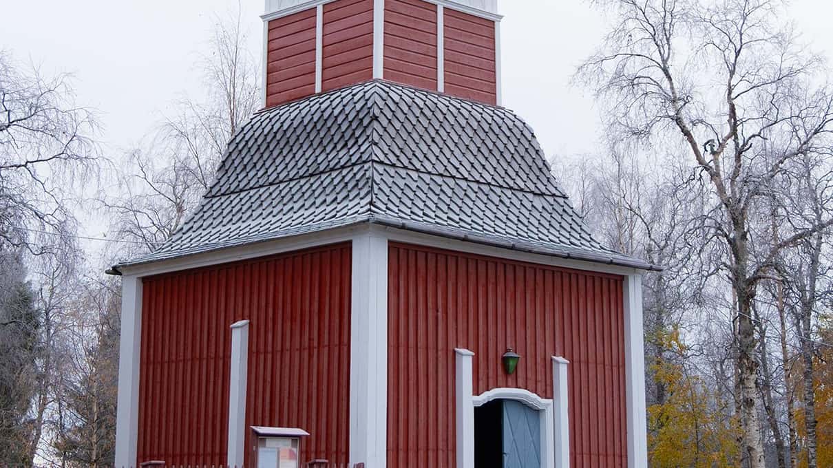 The red wooden bell tower of the Jukkasjärvi church with its shingled roof under snow