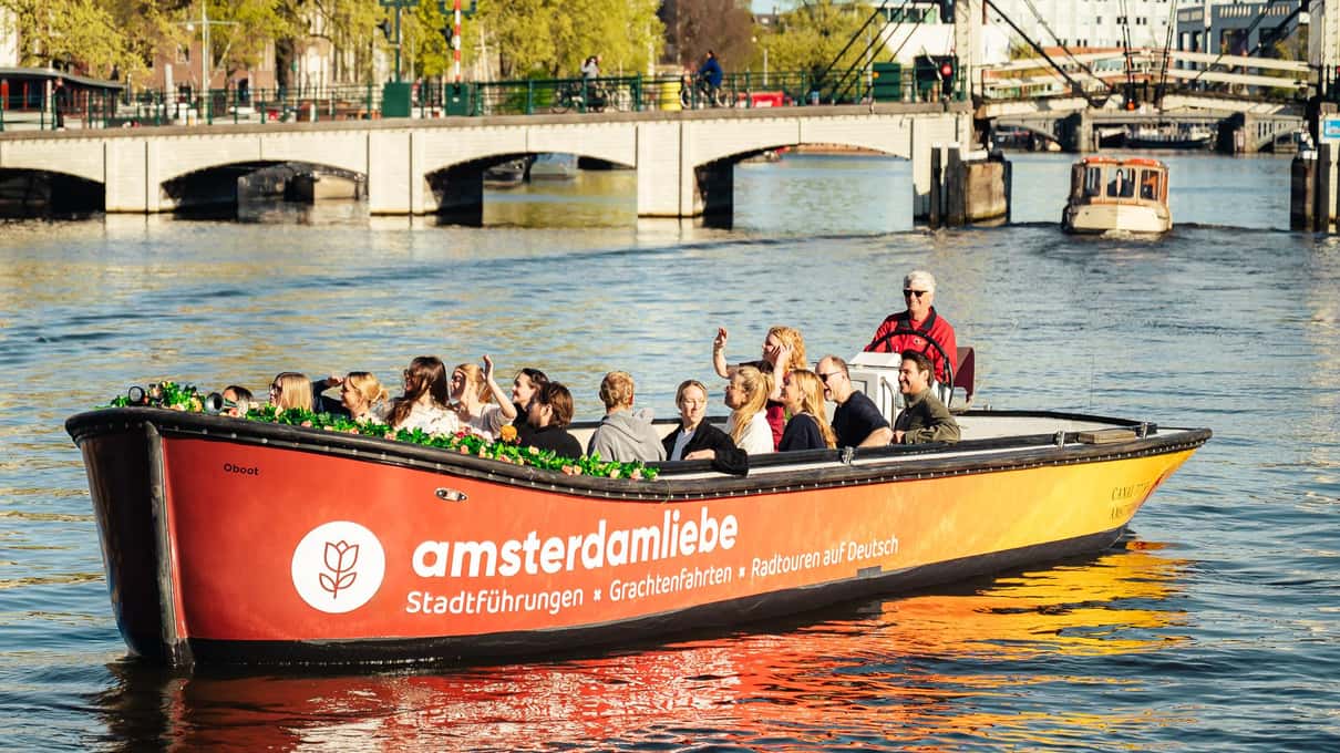 Rotes offenes Boot von Amsterdamliebe voller Gäste auf einer Gracht, im Hintergrund eine Steinbrücke mit Bögen