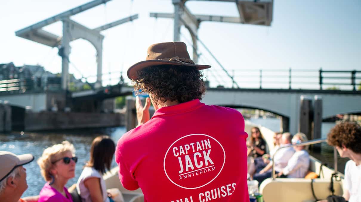 Capitano con la maglietta rosa di Captain Jack e cappello, di spalle, davanti ai passeggeri seduti sulla barca aperta, sullo sfondo la Magere Brug