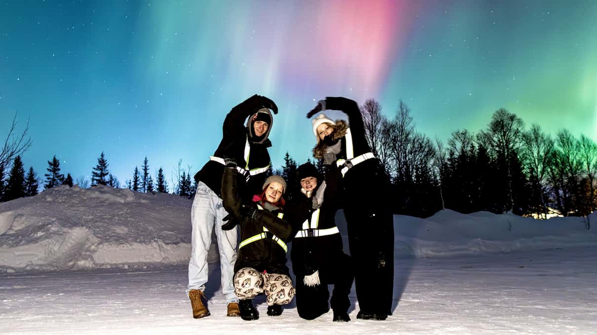 Una familia de cuatro con trajes térmicos negros posando sobre la nieve bajo una aurora rosa y verde, con abetos detrás