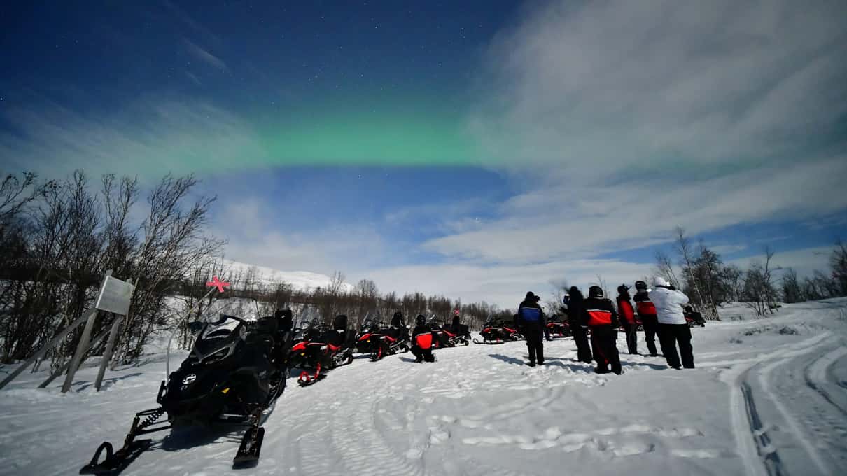 A line of snowmobiles and riders in red suits on a snowy trail at blue dusk under a faint aurora
