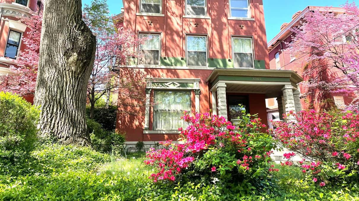 A red-brick Old Louisville mansion behind bright pink azaleas and trees in spring blossom