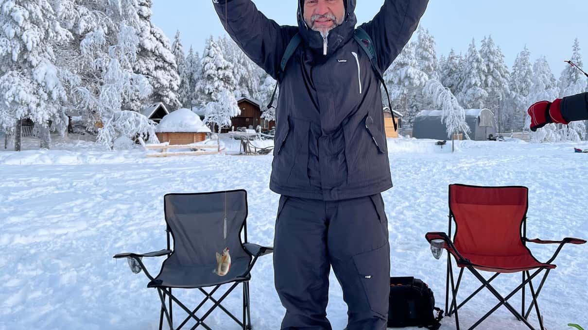 A man in a thick winter jacket raising his arms on the snow beside two camping chairs
