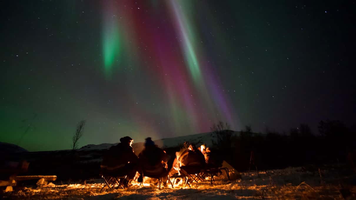 Green and pink Northern Lights rippling over a small group gathered around a bonfire in the snow