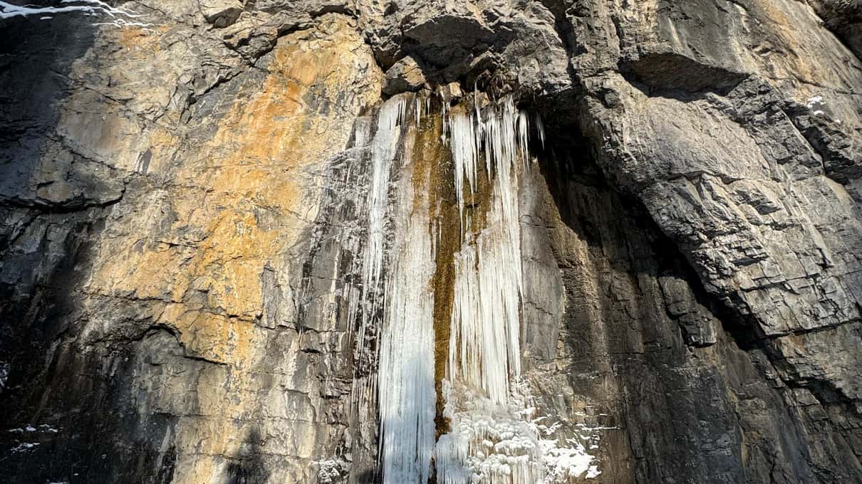 A winter boot with a chain-and-spike ice cleat strapped on, standing in front of a frozen waterfall