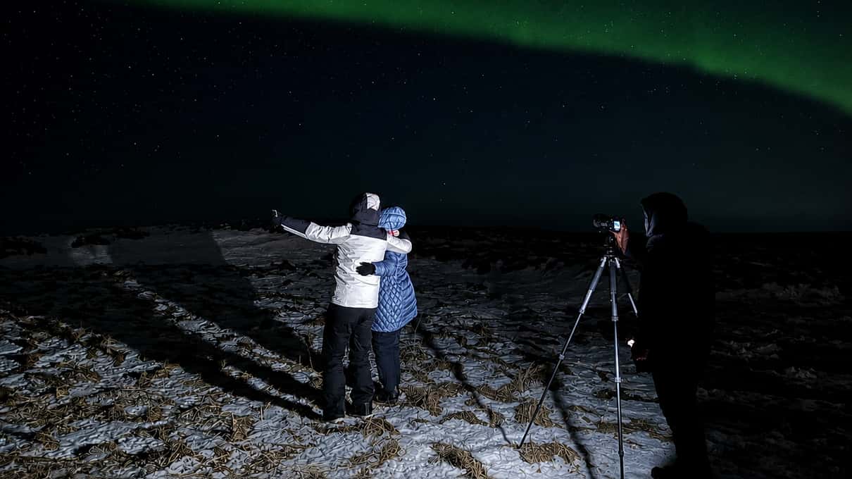 A guest with arms spread wide on snowy ground beside a camera tripod under a green aurora