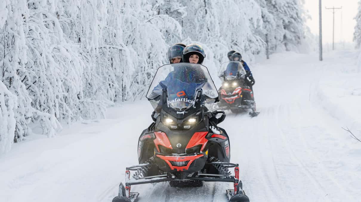 Snowmobiles with headlights on driving along a snow-covered forest track lined with frosted trees