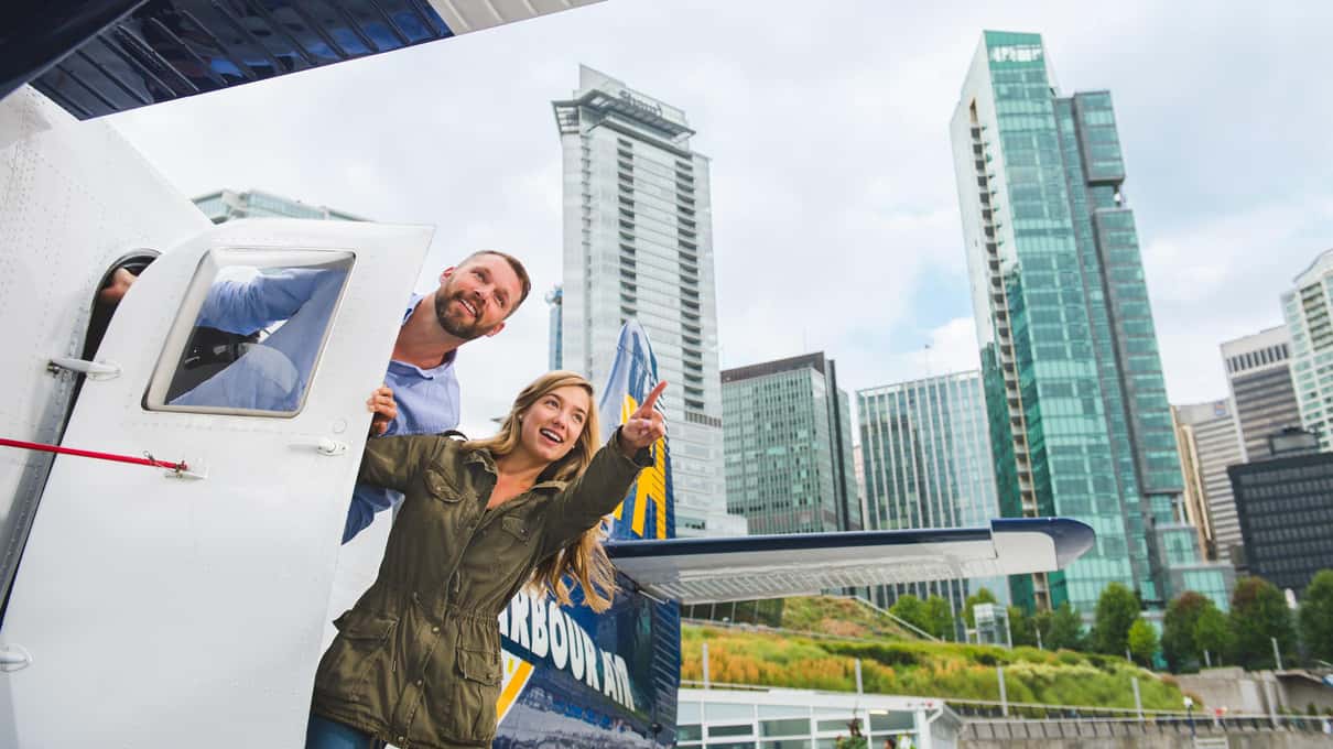 Smiling couple leaning out of the open door of a floatplane with downtown towers behind them