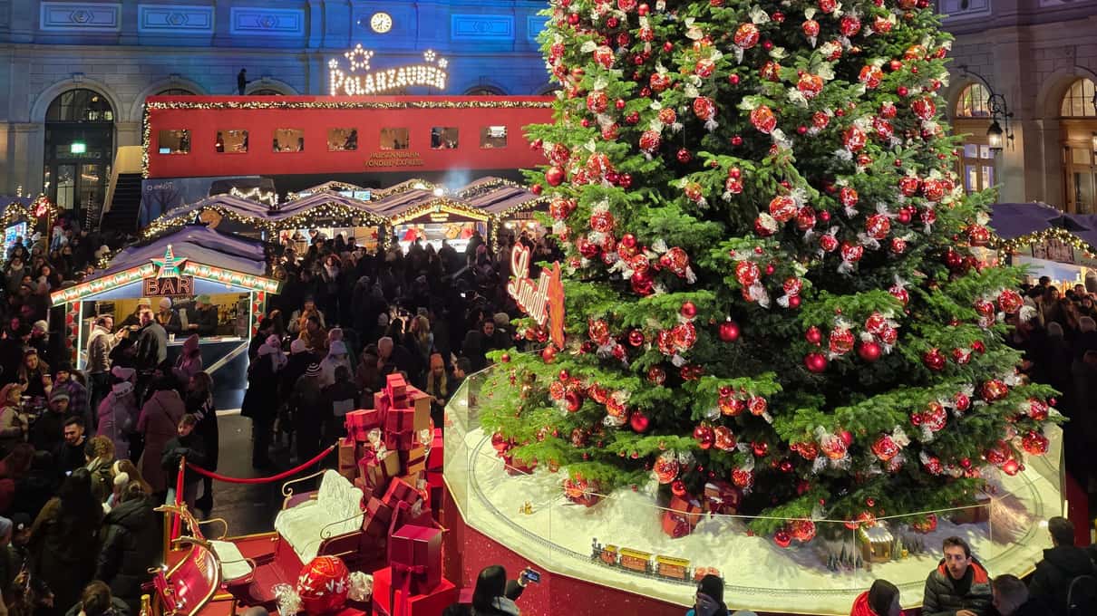 A tall Christmas tree hung with red ornaments in the hall of Zurich main station, with crowds and market huts around it
