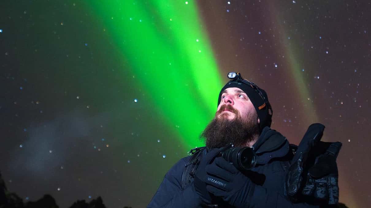 A bearded photographer wearing a headlamp holds a camera and looks up at a green aurora