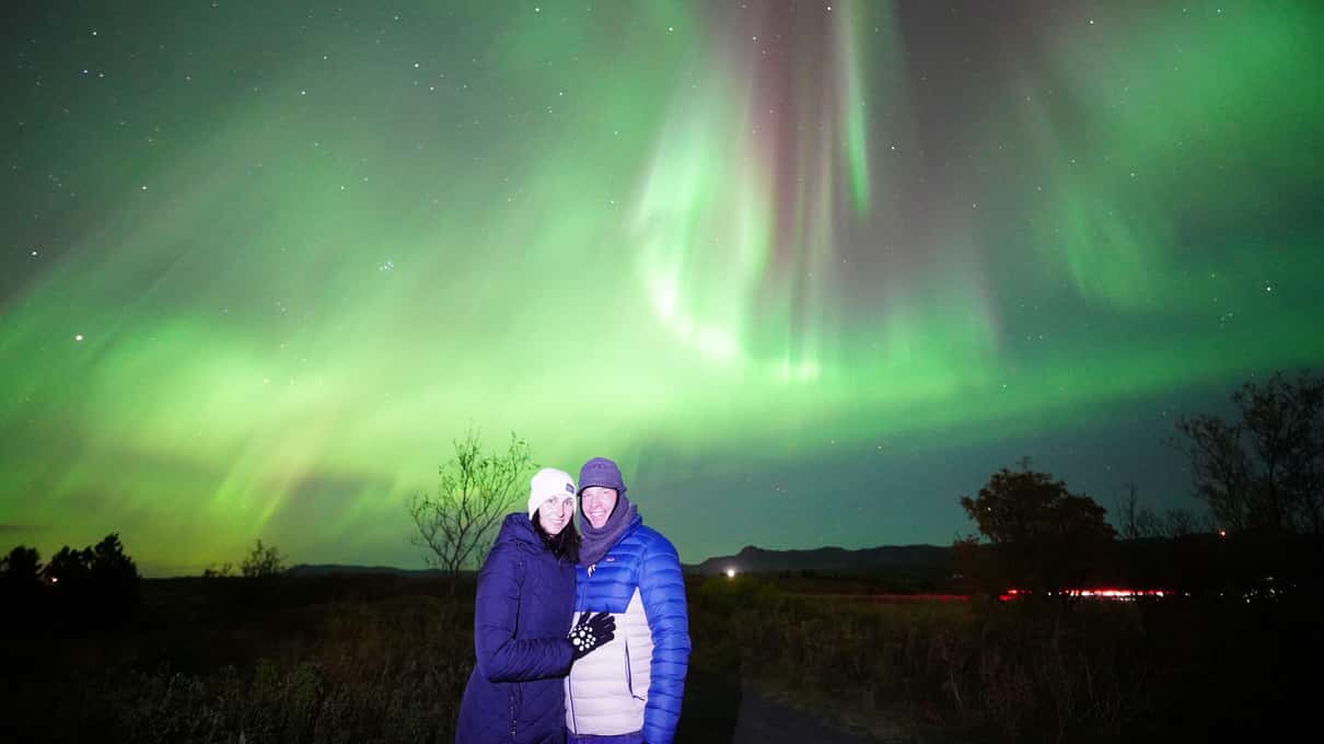 Two people in winter coats standing under a tall green aurora curtain on a clear night near Reykjavík