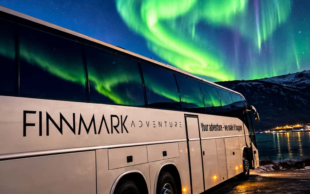 A white Finnmark Adventure coach parked on a snowy road under a band of green aurora