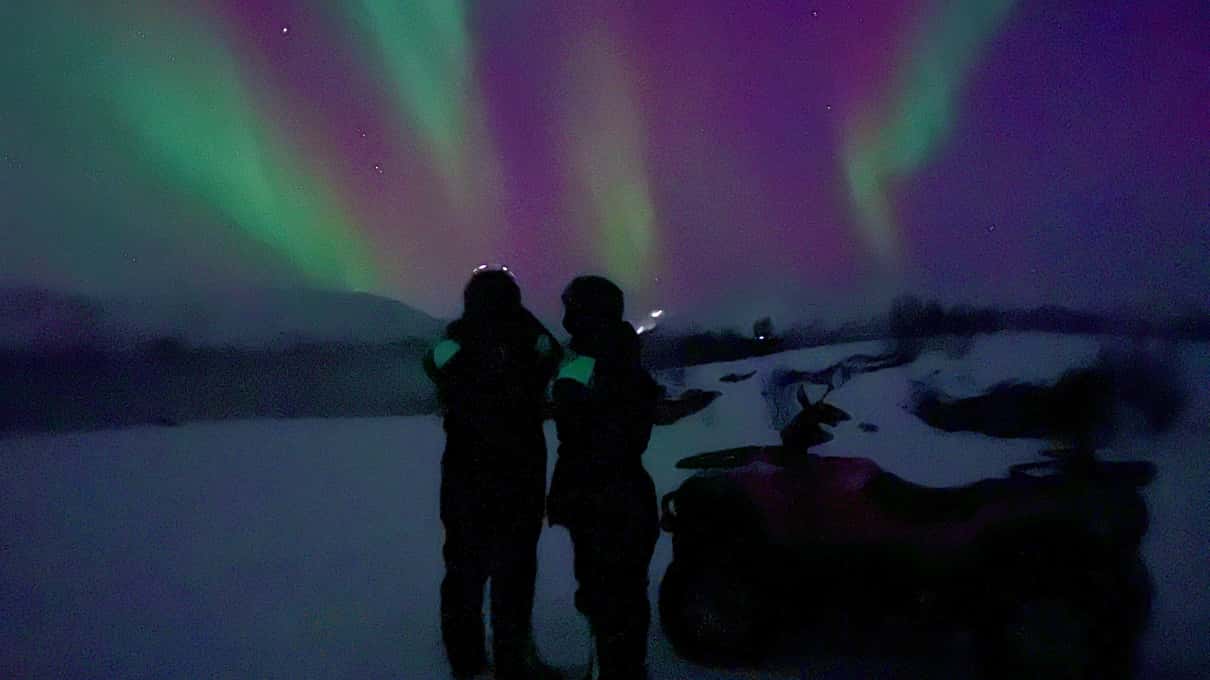Two people standing beside a quad bike under a violet and green aurora on a dark shore