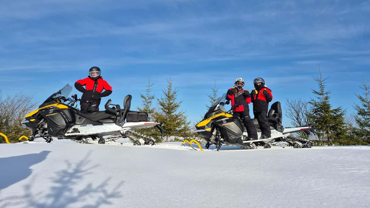 Riders in red jackets standing by two snowmobiles on a snowy ridge under a blue sky