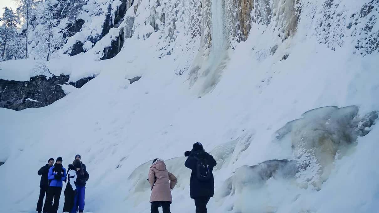 Hikers in winter jackets looking up at a tall white ice fall inside Korouoma canyon