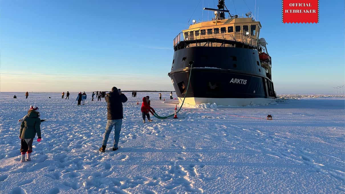 Guests in thick winter clothes walking on the sea ice in front of the black bow of the icebreaker Arktis