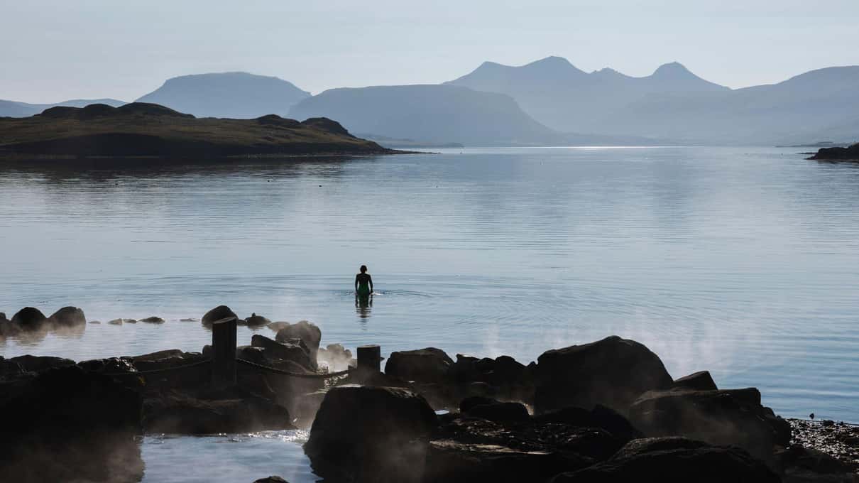A lone bather standing in the calm sea off Hvammsvik with mountains across the fjord