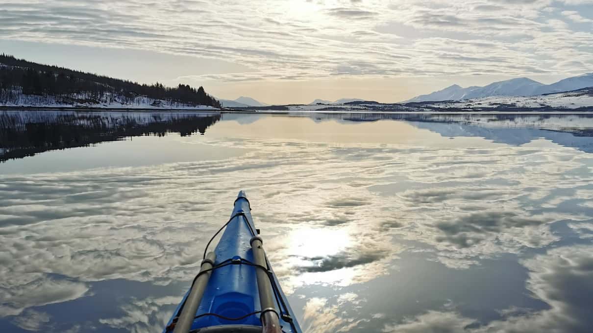 The bow of a blue sea kayak resting on a mirror-calm bay with clouds reflected in the water
