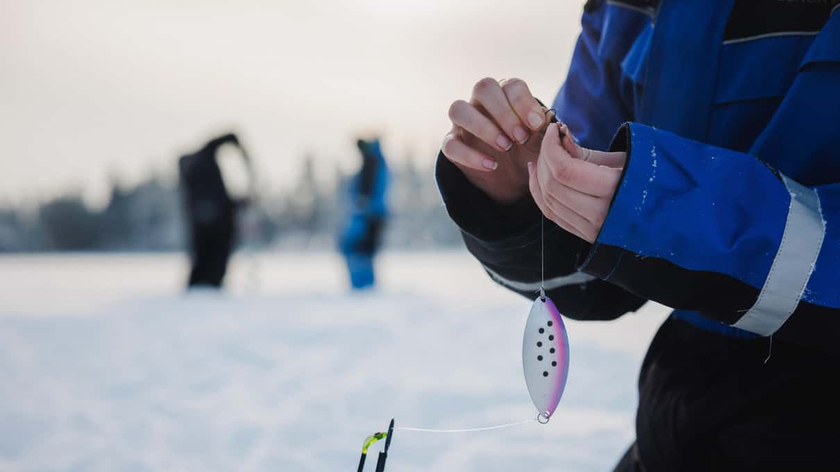 Gloved hands holding a small spotted ice fishing lure above the snow on a frozen lake