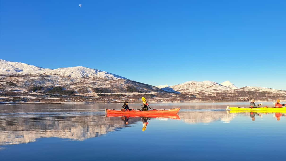 Red and yellow sea kayaks on a mirror-calm bay under snow-covered mountains and a blue sky