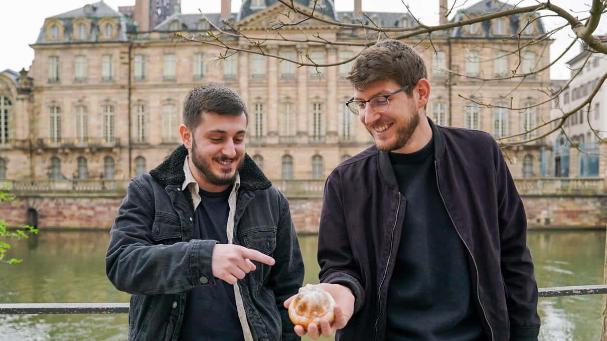Two young men sharing a soft bretzel on a riverside walk, with a grand classical palace across the water behind them