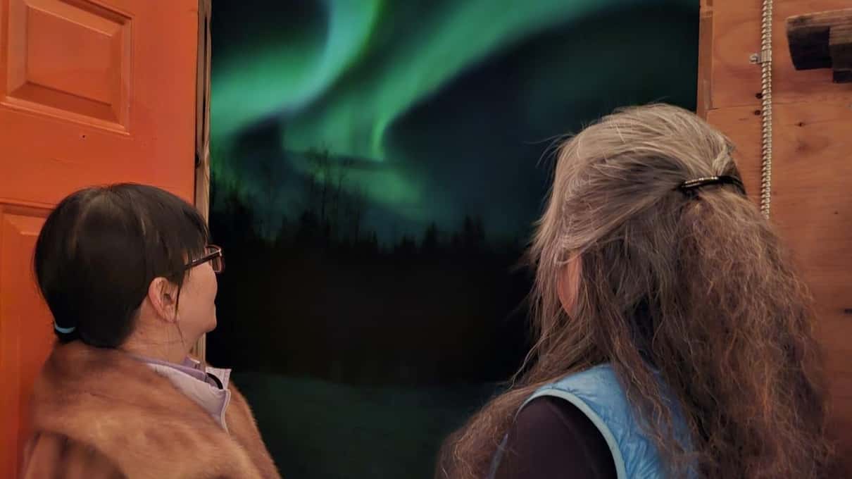 Two guests at the door of a warm cabin looking out at a green aurora over the trees