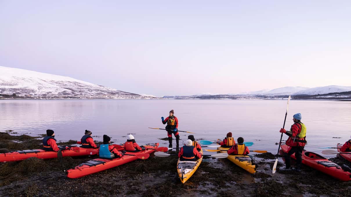 A guide briefing a group of kayakers in red drysuits on a rocky shore before launching
