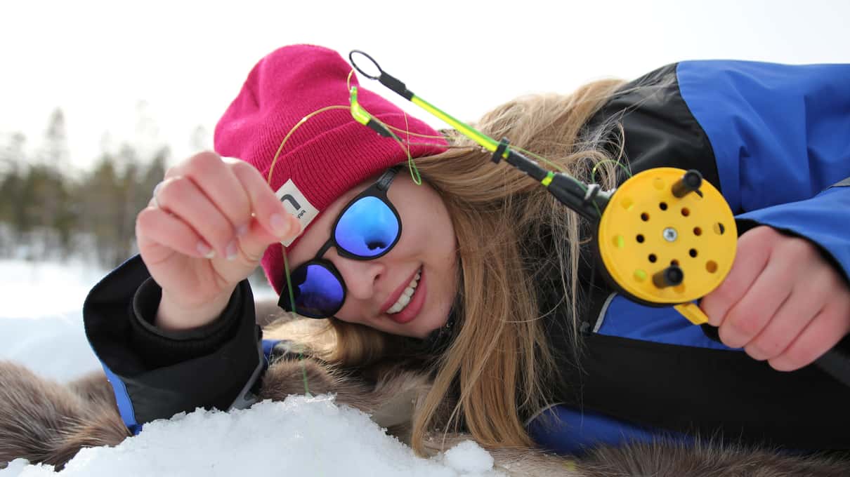 A smiling guest in a pink hat and blue overall lying on the snow beside an ice hole with a short rod
