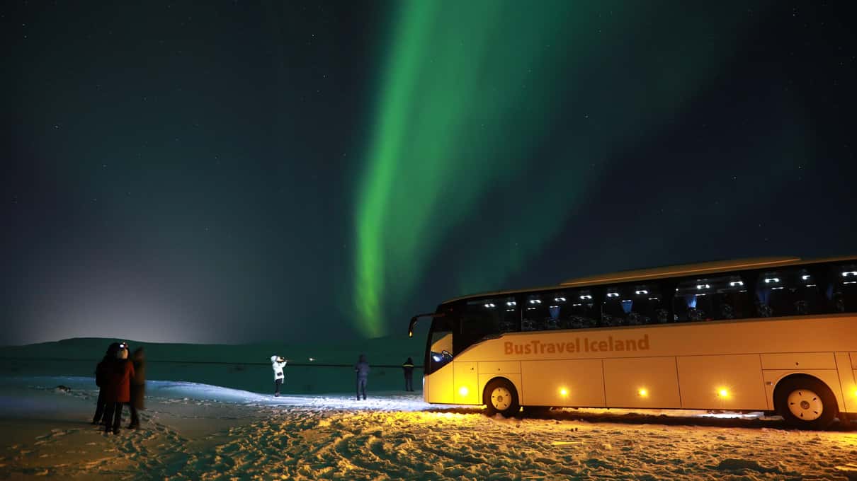 Beleuchteter Reisebus der Nordlichter-Tour auf einer Schneefläche in Island unter grünem Polarlicht