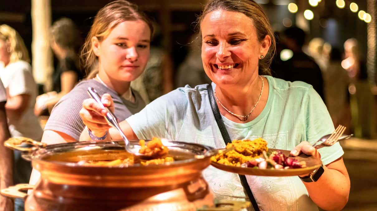 Dos viajeras sonrientes se sirven arroz y guisos de una gran fuente en el bufé nocturno del campamento