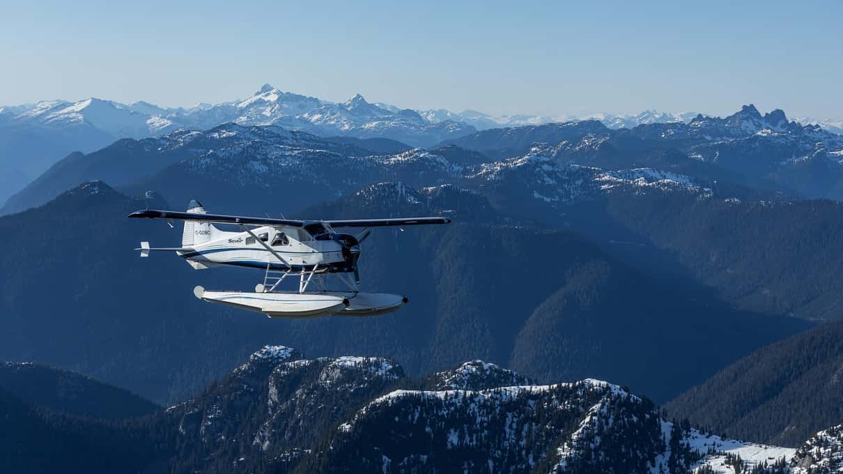 A Beaver floatplane flying above row after row of snow-capped Coast Mountain peaks under a clear blue sky