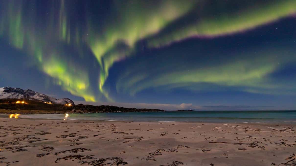 A green aurora arc curving over a pale sandy beach with snowy mountains on the horizon