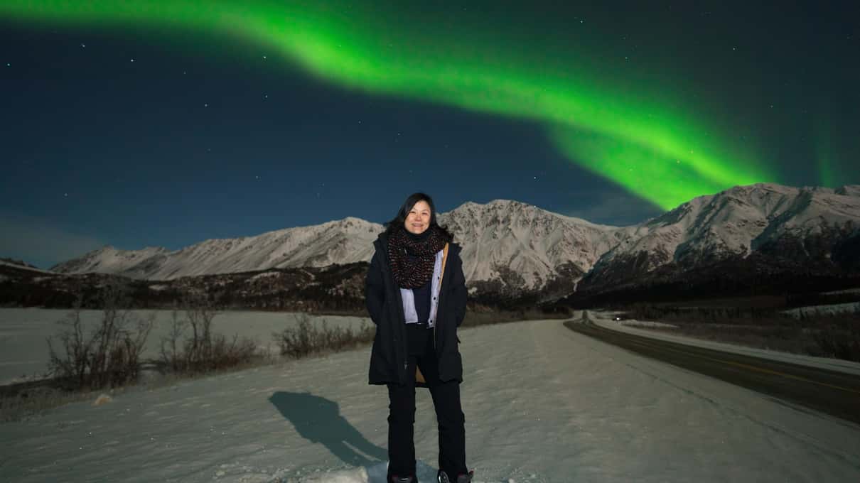 一位游客站在雪路上，身后是黑色的群山和绿色极光