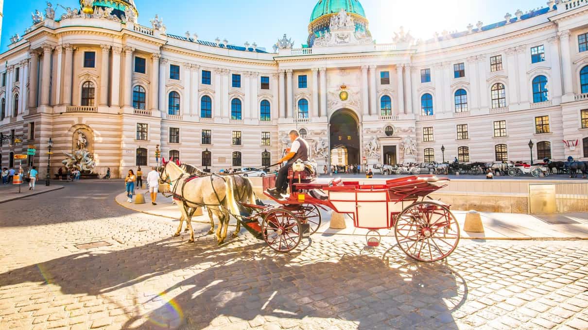 The curved facade of the Hofburg on Michaelerplatz with a Fiaker carriage waiting in front under a blue sky