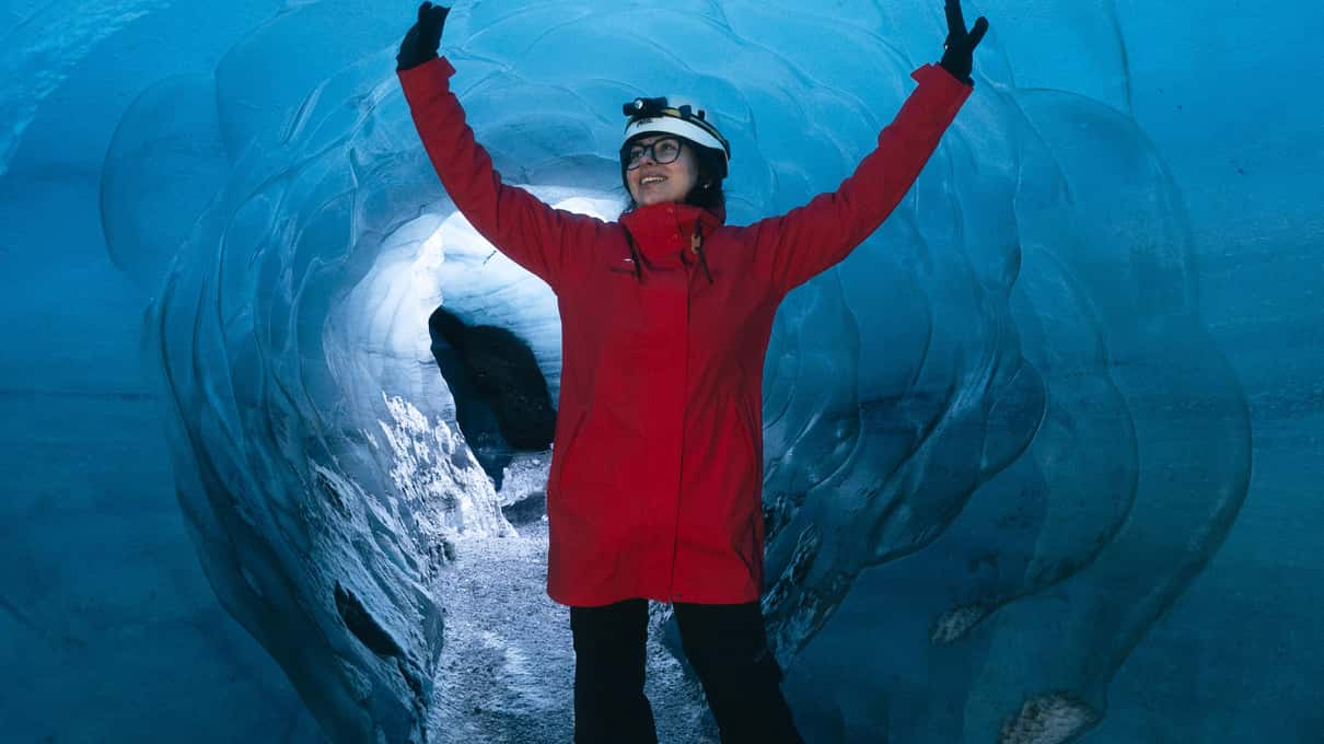 Un visiteur en veste rouge et casque, les deux bras levés dans un tunnel de glace d'un bleu profond