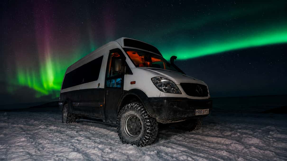 A white high-roofed 4x4 minibus parked on snowy ground under a green and violet aurora