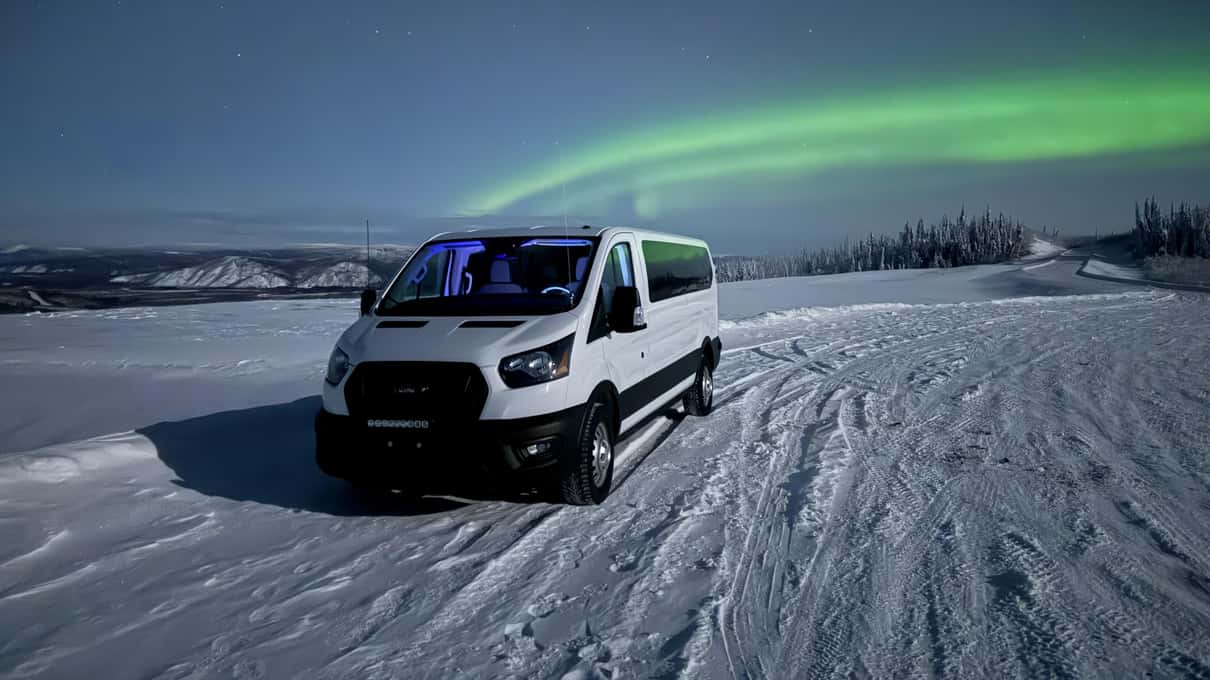 一辆白色旅游车行驶在积雪的公路上，头顶是绿色极光