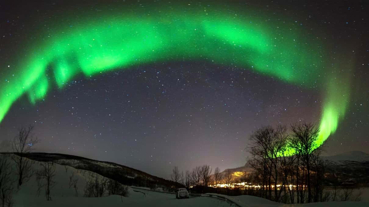 Aurora verde formando un arco sobre colinas nevadas y árboles sin hojas, con las luces de una casa abajo
