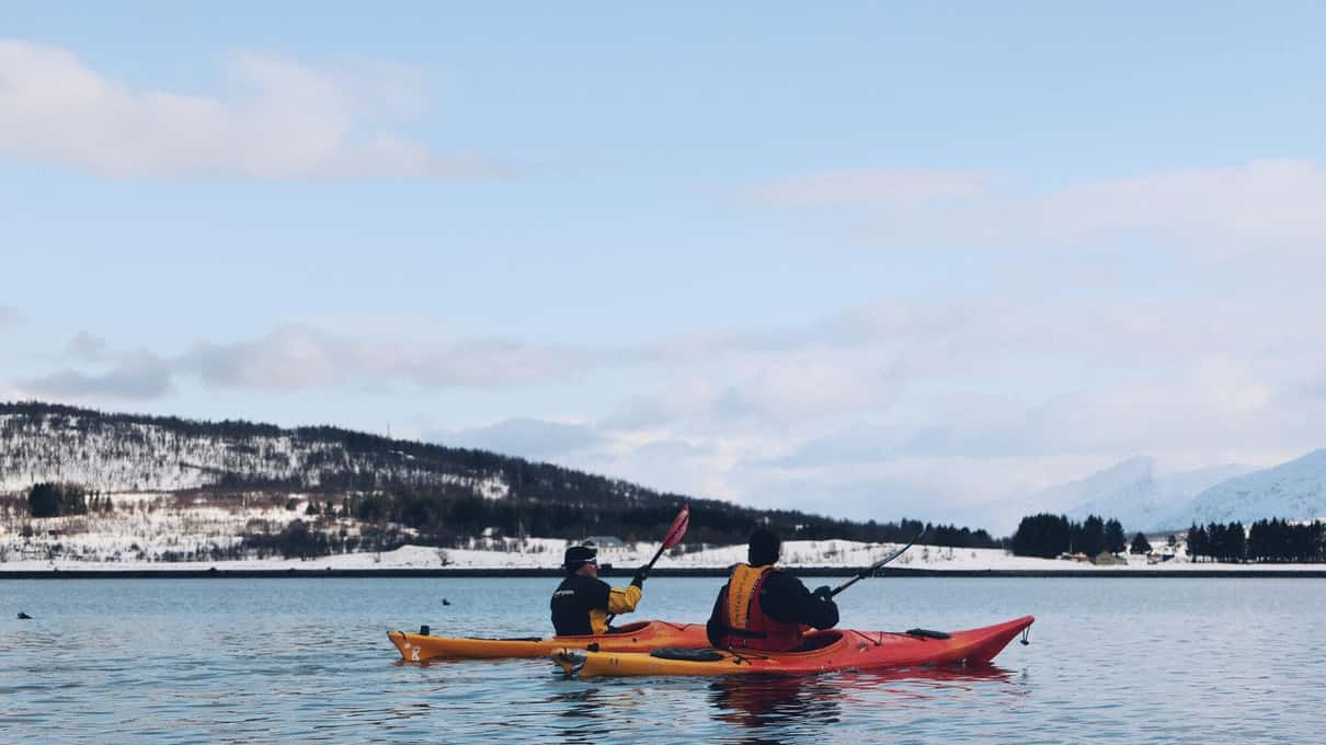 Two paddlers sharing a red and orange double sea kayak near a snowy Tromsø shore