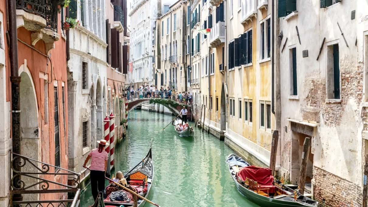 Canal estrecho y tranquilo entre casas blancas y ocres, con barcas rojas amarradas y un puente al fondo