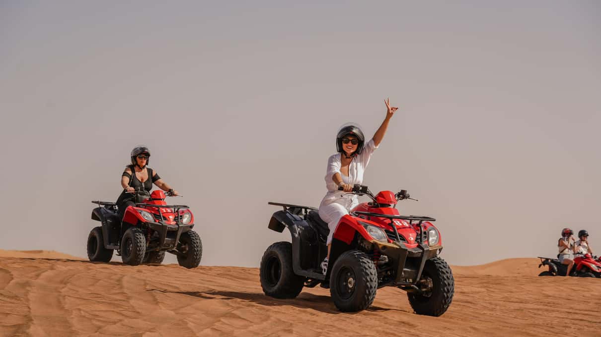 Due persone con il casco guidano quad rossi sulla sabbia, una alza il braccio per salutare