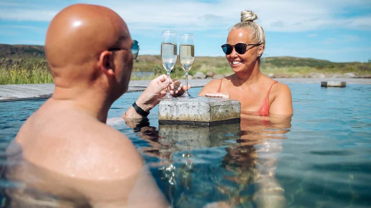 Two bathers raising glasses in a hot pool at the Vök Baths with the lake behind them
