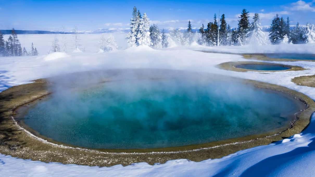 A deep blue hot pool steaming in the snow with frosted trees on the far side