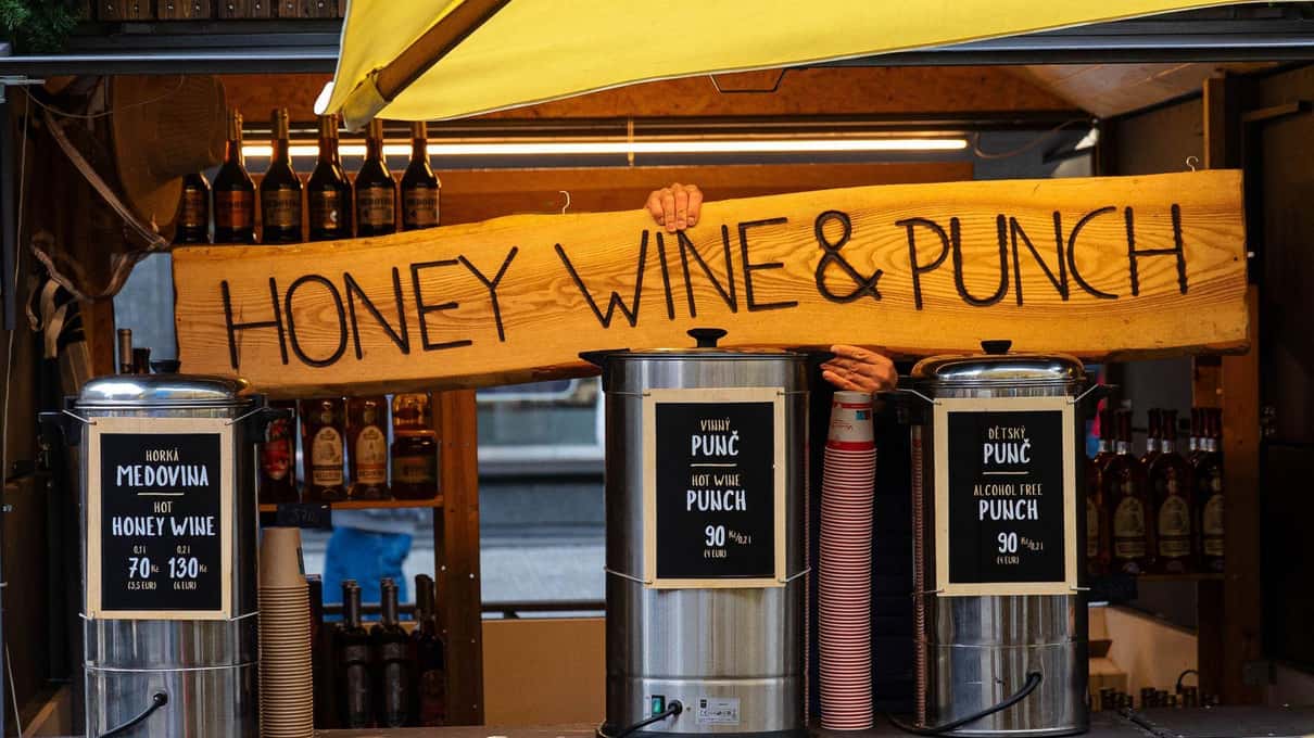 A wooden Christmas market stall in Prague with a hand-painted sign for honey wine and punch and steaming drink urns on the counter