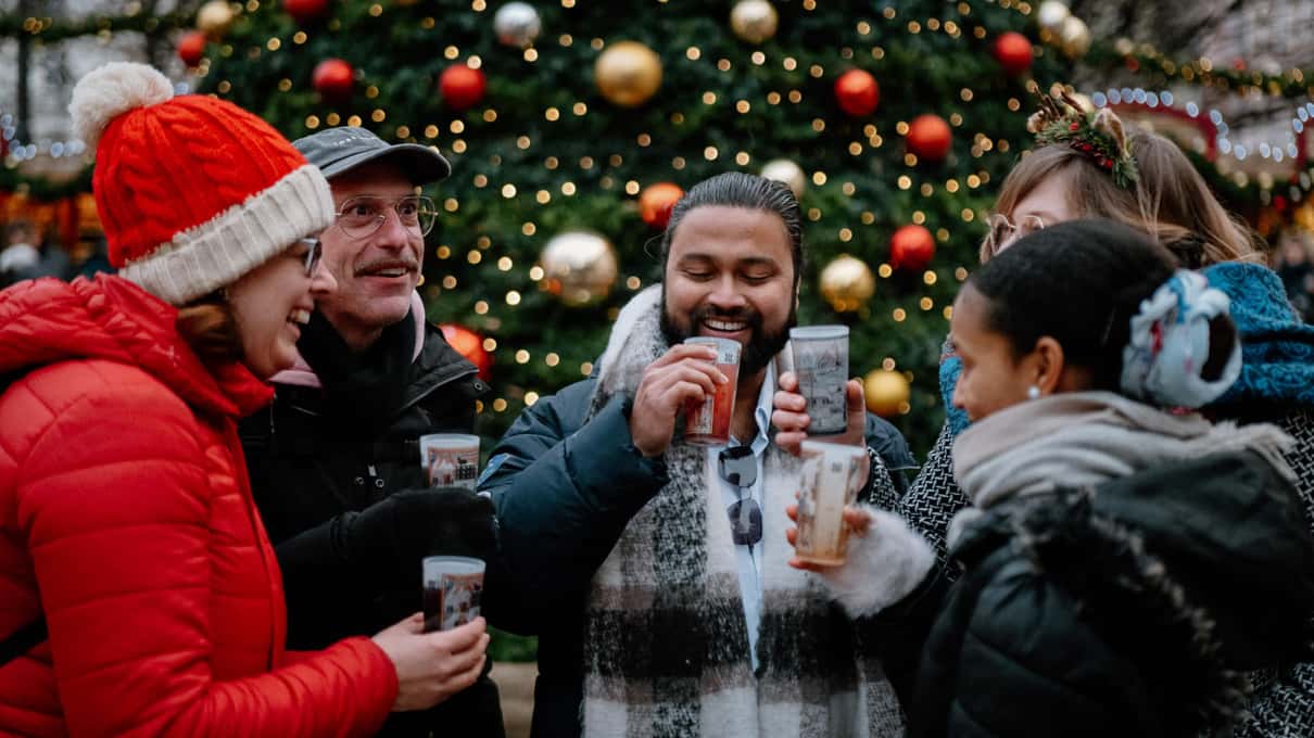 A small group of friends holding warm mugs together under a big lit Christmas tree in Prague