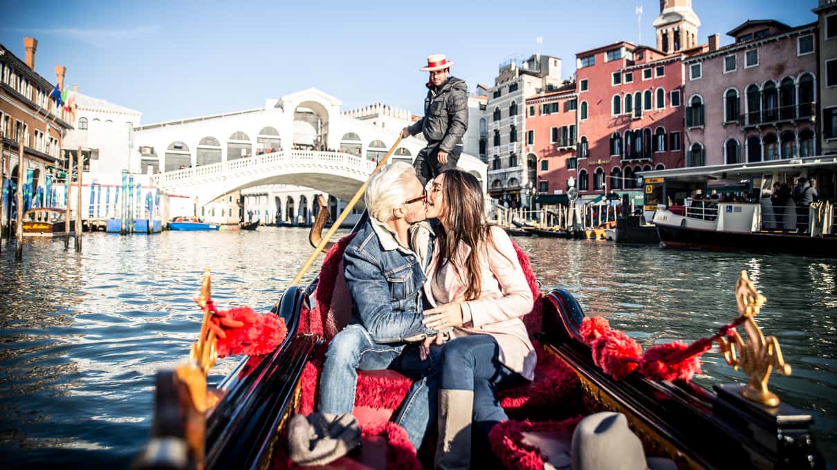 Paar küsst sich in einer Gondel mit roten Polstern auf dem Canal Grande, dahinter die Rialtobrücke und der Gondoliere