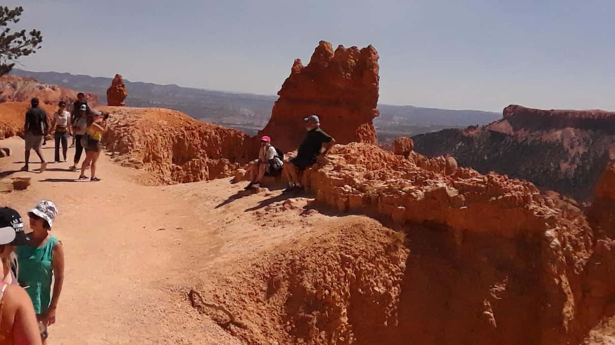 Besucher auf dem Rim Trail im Bryce Canyon neben einem hohen Hoodoo, einige sitzen auf dem Felsvorsprung mit dem Amphitheater im Rücken